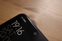 A close-up view of the top corner of a smartphone, displaying the time 19:16, the day SAT, and the date 20/10. The phone screen shows two network providers, Jio and BSNL, in the top right corner. The phone is placed on a wooden surface, with a smooth and slightly reflective finish.