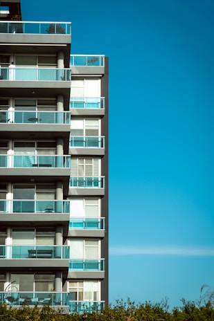 A modern apartment building with multiple floors featuring glass balconies and large windows against a clear blue sky.