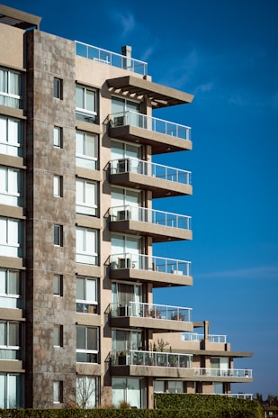 A modern, multi-story apartment building with large balconies and glass railings against a clear blue sky. The structure features a mix of stone and concrete with multiple windows.