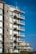 A modern, multi-story apartment building with large balconies and glass railings against a clear blue sky. The structure features a mix of stone and concrete with multiple windows.