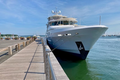 A large white yacht is docked beside a wooden pier with clear blue skies overhead. The pier extends alongside the water, with potted plants and benches lining the walkway. Several smaller boats are visible in the background.
