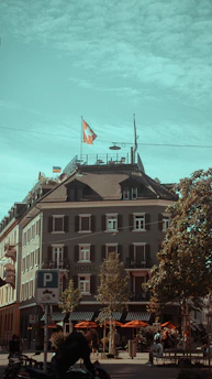 A multi-story building with a Swiss flag on the roof, possibly a hotel, is located at the corner of a street. The facade is adorned with several windows featuring shutters. Outdoor seating with orange umbrellas is set up on the ground floor. Trees line the street, and there are a few people walking or biking nearby. A parking sign for bicycles is visible in the foreground.