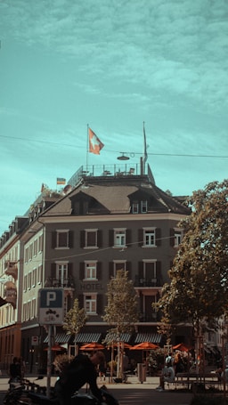A multi-story building with a Swiss flag on the roof, possibly a hotel, is located at the corner of a street. The facade is adorned with several windows featuring shutters. Outdoor seating with orange umbrellas is set up on the ground floor. Trees line the street, and there are a few people walking or biking nearby. A parking sign for bicycles is visible in the foreground.
