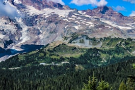 A stunning mountainous landscape featuring rugged peaks topped with patches of snow and ice. Below the mountains, lush green forests extend across rolling hills, creating a serene and majestic natural scene under a clear blue sky with scattered clouds.