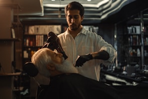 A barber is styling a customer's hair in a dimly lit barbershop. The focus is on the barber who is wearing a white shirt and black gloves, using a hairdryer on the customer's white beard. Shelves with various hair products and a stylish black ceiling are visible in the background.