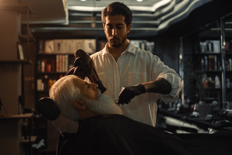 A barber is styling a customer's hair in a dimly lit barbershop. The focus is on the barber who is wearing a white shirt and black gloves, using a hairdryer on the customer's white beard. Shelves with various hair products and a stylish black ceiling are visible in the background.