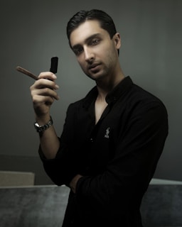 A man with short dark hair and a neat beard is holding a straight razor. He is wearing a dark buttoned shirt with a logo on the chest. His pose appears confident and slightly relaxed, with an arm crossed over his body. The background is a muted green, adding to the overall composed and intimate atmosphere.