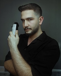 A man with short hair and a trimmed beard holds an electric hair clipper close to his face. He is wearing a dark shirt and has a serious expression, looking directly at the camera. The background is a muted gray-green color, creating a calm and focused atmosphere.