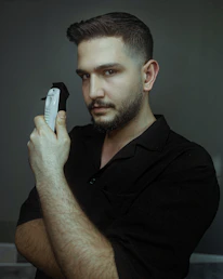 A man with short hair and a trimmed beard holds an electric hair clipper close to his face. He is wearing a dark shirt and has a serious expression, looking directly at the camera. The background is a muted gray-green color, creating a calm and focused atmosphere.