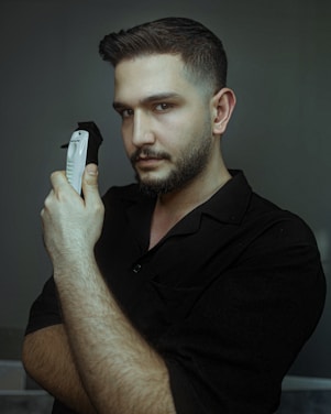 A man with short hair and a trimmed beard holds an electric hair clipper close to his face. He is wearing a dark shirt and has a serious expression, looking directly at the camera. The background is a muted gray-green color, creating a calm and focused atmosphere.
