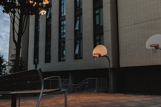 A quiet urban scene featuring a basketball court in front of a large building with tall windows. The setting appears to be during dusk or early evening, with streetlights casting a soft glow over the area. A bench and a tree are visible, adding to the serene atmosphere.