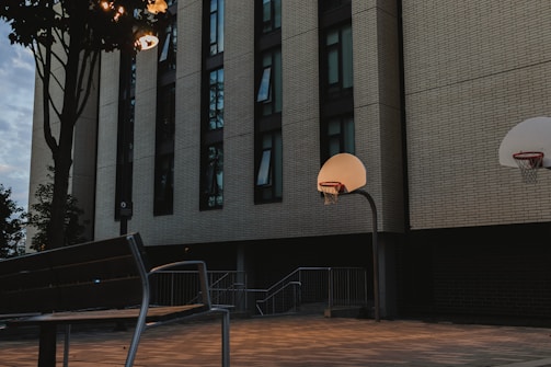 A quiet urban scene featuring a basketball court in front of a large building with tall windows. The setting appears to be during dusk or early evening, with streetlights casting a soft glow over the area. A bench and a tree are visible, adding to the serene atmosphere.
