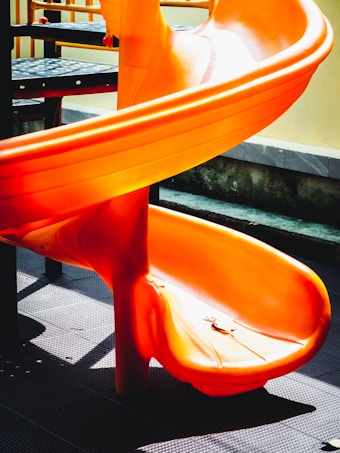 A bright orange spiral slide on a playground, casting shadows on the textured black surface beneath. The structure appears vibrant and inviting, with sunlight emphasizing the curves and smooth surface of the slide.