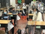 Several students wearing masks are seated at desks in a classroom, which are separated by clear plastic barriers. The students appear to be engaged and focused. There are educational materials and a blackboard visible in the background.