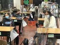 Several students wearing masks are seated at desks in a classroom, which are separated by clear plastic barriers. The students appear to be engaged and focused. There are educational materials and a blackboard visible in the background.