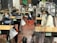Several students wearing masks are seated at desks in a classroom, which are separated by clear plastic barriers. The students appear to be engaged and focused. There are educational materials and a blackboard visible in the background.
