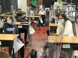 Several students wearing masks are seated at desks in a classroom, which are separated by clear plastic barriers. The students appear to be engaged and focused. There are educational materials and a blackboard visible in the background.