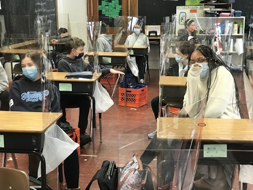 Several students wearing masks are seated at desks in a classroom, which are separated by clear plastic barriers. The students appear to be engaged and focused. There are educational materials and a blackboard visible in the background.