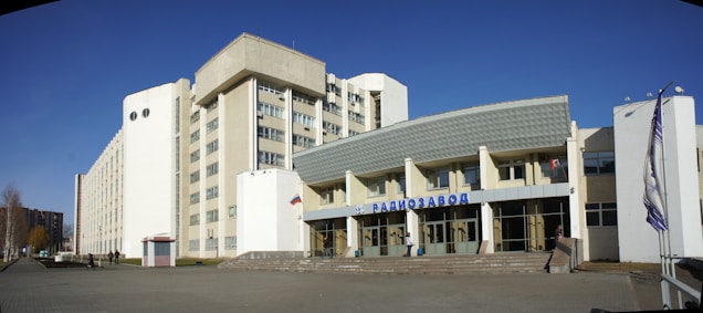 A large building complex with a modern architectural design. The structure has multiple stories, with a white facade and large windows. There is a prominent sign in the front in Cyrillic script, suggesting it could be an industrial or commercial building. Flags are displayed beside the entrance, and the surrounding area is paved and open.