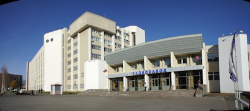 A large building complex with a modern architectural design. The structure has multiple stories, with a white facade and large windows. There is a prominent sign in the front in Cyrillic script, suggesting it could be an industrial or commercial building. Flags are displayed beside the entrance, and the surrounding area is paved and open.
