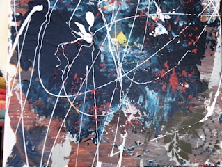An abstract painting featuring dynamic splashes and drips of paint on a dark, textured background. Various colors, including navy blue, white, red, and hints of yellow, blend in chaotic patterns. The white lines create a web-like structure, while the textured surface adds depth and complexity.
