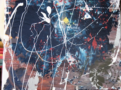 An abstract painting featuring dynamic splashes and drips of paint on a dark, textured background. Various colors, including navy blue, white, red, and hints of yellow, blend in chaotic patterns. The white lines create a web-like structure, while the textured surface adds depth and complexity.