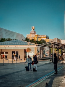 A cityscape with a notable building featuring a large rooftop logo. People are visible in the foreground, some with luggage, suggesting a travel or transit area. The sky is clear and blue, with sunlight casting shadows on the ground. There are modern structures and greenery in the background.