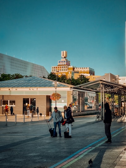 A cityscape with a notable building featuring a large rooftop logo. People are visible in the foreground, some with luggage, suggesting a travel or transit area. The sky is clear and blue, with sunlight casting shadows on the ground. There are modern structures and greenery in the background.