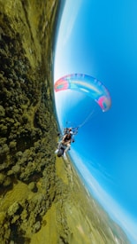 A person is flying in a powered paraglider high above a vast landscape of green forests and fields. The paraglider's colorful wing contrasts against the clear blue sky, offering a panoramic view of the scenery below.