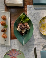 A table setting featuring a green leaf-shaped plate with food topped with a dark sauce and garnished with greens, placed on a patterned blue and white tablecloth. To the left, there is a rectangular white plate with two small food portions. On the right, a decorative round plate with a floral design is partly visible. A stack of napkins is at the top, and a knife lies on the table.