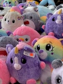 A collection of plush toys with various colors and designs, including unicorns, penguins, and other adorable animals, mostly in pastel and bright hues. The toys are densely packed together, creating a fluffy and vibrant scene.