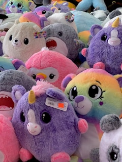 A collection of plush toys with various colors and designs, including unicorns, penguins, and other adorable animals, mostly in pastel and bright hues. The toys are densely packed together, creating a fluffy and vibrant scene.