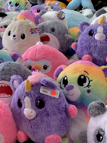 A collection of plush toys with various colors and designs, including unicorns, penguins, and other adorable animals, mostly in pastel and bright hues. The toys are densely packed together, creating a fluffy and vibrant scene.