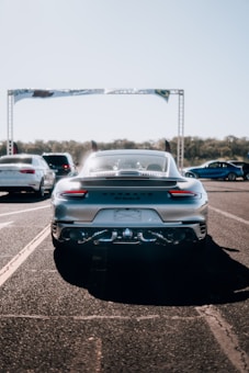 Several cars are positioned on a road, with a prominent focus on a sleek, silver sports car in the foreground. This car's license plate area is blurred, and it appears to be parked on a track or runway. In the background, more vehicles can be seen along with a large banner overhead.