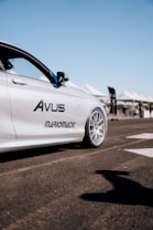 A sleek, silver car with distinctive branding is parked on a paved surface, with a clear blue sky in the background. There are white tents and flags, suggesting the setting is an outdoor event or exhibition.