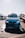 A blue BMW car is positioned on a transport trailer, with its headlights glowing. The background features other vehicles, including a truck to the left and another car on a trailer to the right. The sun is shining brightly, casting reflections on the car's surface.