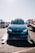 A blue BMW car is positioned on a transport trailer, with its headlights glowing. The background features other vehicles, including a truck to the left and another car on a trailer to the right. The sun is shining brightly, casting reflections on the car's surface.