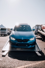 A blue BMW car is positioned on a transport trailer, with its headlights glowing. The background features other vehicles, including a truck to the left and another car on a trailer to the right. The sun is shining brightly, casting reflections on the car's surface.