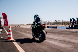 A motorcyclist wearing protective gear rides a sport motorcycle on an asphalt racing track. There are orange traffic cones and multiple red banners with logos along the track. In the background, spectators and more banners are visible, as well as a clear blue sky.