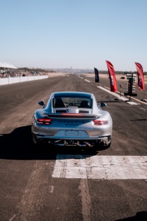 A sleek silver sports car is positioned on a wide, open race track. The vehicle appears to be a Porsche 911 Turbo S, seen from behind, ready to speed down the straight path. Bright red flags and a timing light tower are situated along the side of the track, with spectators gathered in the distance under a clear blue sky.