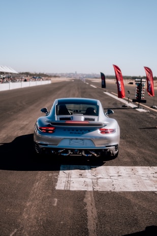 A sleek silver sports car is positioned on a wide, open race track. The vehicle appears to be a Porsche 911 Turbo S, seen from behind, ready to speed down the straight path. Bright red flags and a timing light tower are situated along the side of the track, with spectators gathered in the distance under a clear blue sky.