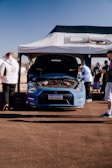 A high-performance car with its hood open is displayed under a canopy with branding. Several people are gathered around, examining the car. The scene is set outdoors under a clear blue sky, suggesting a car show or exhibition.