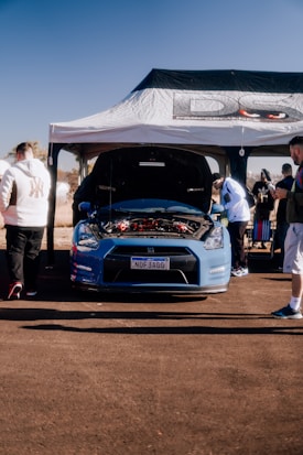 A high-performance car with its hood open is displayed under a canopy with branding. Several people are gathered around, examining the car. The scene is set outdoors under a clear blue sky, suggesting a car show or exhibition.