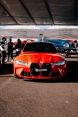 A bright red sports car with a sleek design is parked under a tent. The car is positioned centrally, showcasing its aggressive front grille and stylish headlights. Several people are gathered in the background, some appearing to converse.