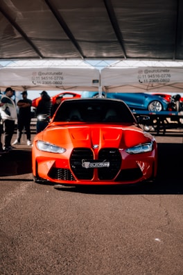A bright red sports car with a sleek design is parked under a tent. The car is positioned centrally, showcasing its aggressive front grille and stylish headlights. Several people are gathered in the background, some appearing to converse.