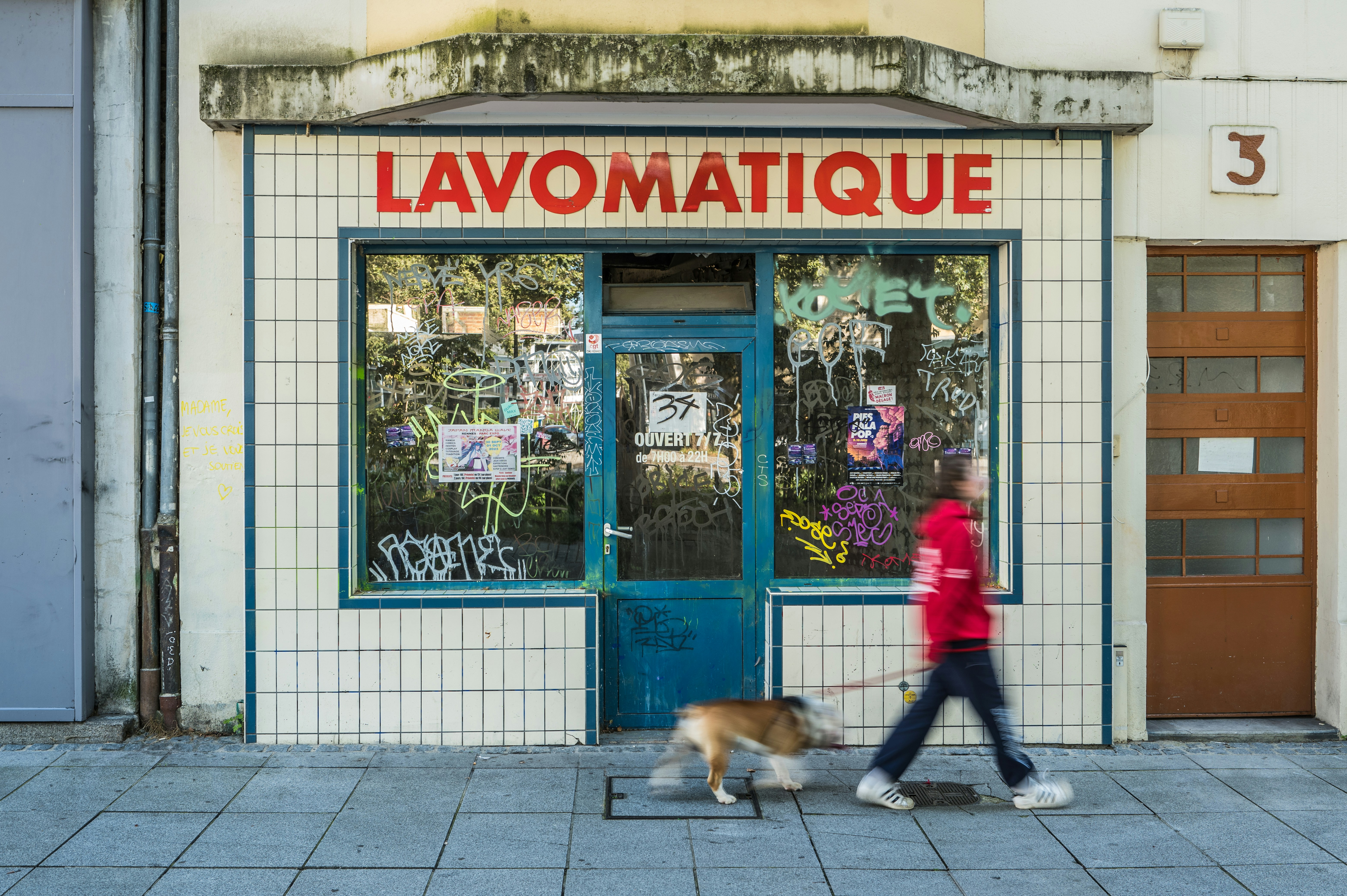 Former laundromat (“Lavomatique”) located on Place de Bretagne in Rennes, France. The storefront is covered with graffiti and stickers, while a person walks by with a dog in motion blur. Captured with a Leica Q3.