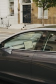 A modern car is parked on the street with a lit screen visible inside, displaying a map. The car is positioned in front of a brick building with a black door and white trim. A wrought iron fence lines the sidewalk, and a tree is present next to the car.