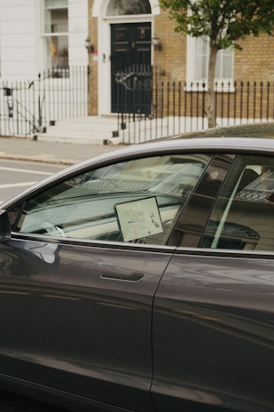 A modern car is parked on the street with a lit screen visible inside, displaying a map. The car is positioned in front of a brick building with a black door and white trim. A wrought iron fence lines the sidewalk, and a tree is present next to the car.