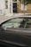 A modern car is parked on the street with a lit screen visible inside, displaying a map. The car is positioned in front of a brick building with a black door and white trim. A wrought iron fence lines the sidewalk, and a tree is present next to the car.