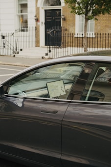 A modern car is parked on the street with a lit screen visible inside, displaying a map. The car is positioned in front of a brick building with a black door and white trim. A wrought iron fence lines the sidewalk, and a tree is present next to the car.